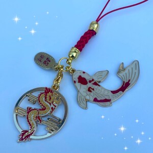Op de afbeelding: Een rode en gouden hanger met een drakenontwerp en een witte en rode koi-vis hanger. De hangers zijn bevestigd aan een rood touwtje met een gouden kraal.