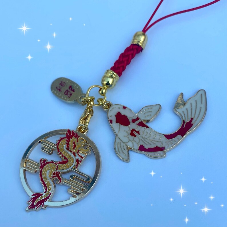 Op de afbeelding: Een rode en gouden hanger met een drakenontwerp en een witte en rode koi-vis hanger. De hangers zijn bevestigd aan een rood touwtje met een gouden kraal.
