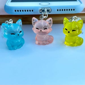 May include: Three translucent fox charms in blue, pink, and yellow. Each charm has a silver-coloured loop for attachment. The foxes are in a sitting position with closed eyes, and are likely phone charms or accessories.