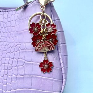 May include: A gold-toned keychain with red floral and fan charms. The charms are attached to a gold ring, hanging against a textured, light purple handbag. The fan charm has a detailed design.