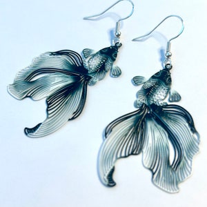 May include: A pair of silver dangle earrings featuring black and white detailed fish designs. The fish have a delicate, intricate design and are a statement piece.