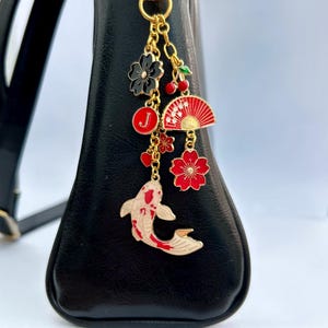 May include: A gold charm featuring a white and red koi fish, a red and gold fan, a red cherry, a black flower, and a red flower. The charm is attached to a gold chain and hangs from a black handbag.
