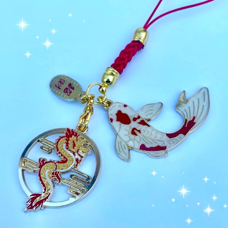 Op de afbeelding: Een gouden bedel met een rood en gouden drakenontwerp, een witte en rode koi-visbedel en een gouden bedel met de tekst "Kyo 22" in rood. De bedels zijn bevestigd aan een rood gevlochten koord.