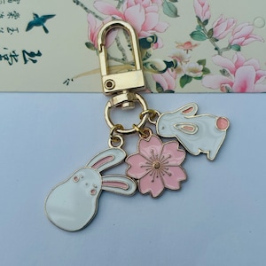 May include: A gold key chain with three charms: two white bunny charms and a pink flower charm. The flower charm has a gold center.