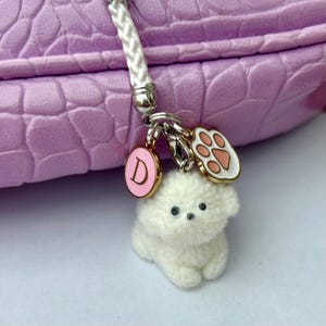 May include: A white fluffy dog charm with a pink paw print charm and a gold charm with the letter D. The charms are attached to a white cord.