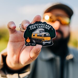 以下が含まれることがあります： キャンピングカーと「Weekends are for camping」というテキスト、焚き火、キャンピングカーの下に「Project Wander」という単語が書かれた黒と黄色のステッカーです。