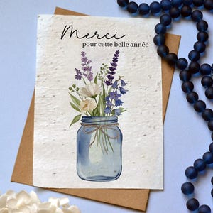 Peut inclure: Une carte de vœux rectangulaire en papier texturé blanc cassé avec une illustration à l'aquarelle d'un bocal en verre bleu rempli de lavande, de campanules et de fleurs blanches. Le texte indique Merci pour cette belle année en noir.