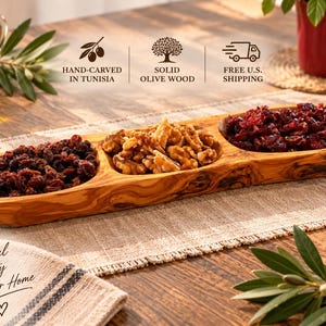 May include: An elongated, three-section olive wood serving tray with a natural grain pattern. The tray contains dried cranberries in the left and right sections, and walnut halves in the center section. Text on the image includes Hand-carved in Tunisia, Solid Olive Wood, Free U.S. Shipping, and Natural Beauty for Your Home.
