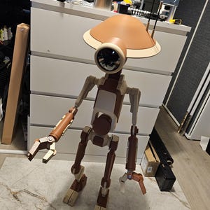 May include: A detailed, life-size robot model with a tan and cream color scheme. The robot has a large, circular eye, a domed head, and articulated limbs. It stands on a light-colored floor, with a cluttered background.