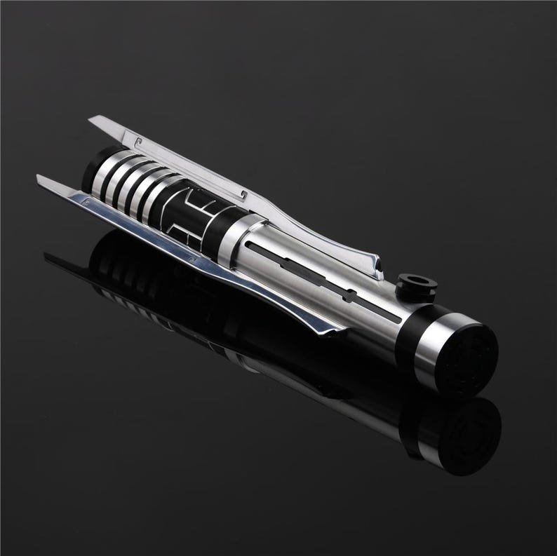 May include: A silver and black lightsaber with a textured handle and a black blade. The lightsaber is on a black surface.