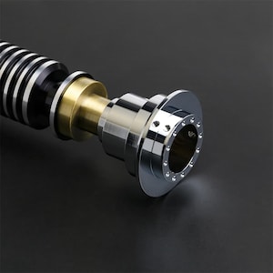 May include: A close-up of a lightsaber hilt with a black and silver body, a gold accent, and a chrome end cap with a ring of small holes.