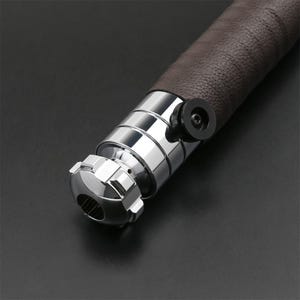 May include: Close-up of a silver lightsaber hilt with a brown leather grip. The hilt has a textured metal design and a black knob.