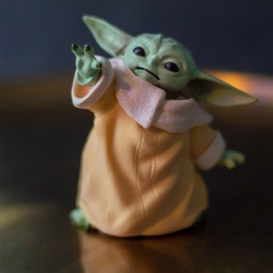 K&ouml;nnte beinhalten: Eine Figur von Grogu, auch bekannt als Baby Yoda, aus The Mandalorian. Die Figur hat gro&szlig;e Ohren, gr&uuml;ne Haut und tr&auml;gt eine beige Robe mit wei&szlig;em Besatz. Die Figur ist mit einer erhobenen Hand posiert.