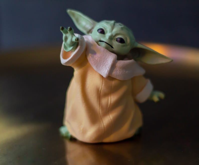 K&ouml;nnte beinhalten: Eine Figur von Grogu, auch bekannt als Baby Yoda, aus The Mandalorian. Die Figur hat gro&szlig;e Ohren, gr&uuml;ne Haut und tr&auml;gt eine beige Robe mit wei&szlig;em Besatz. Die Figur ist mit einer erhobenen Hand posiert.