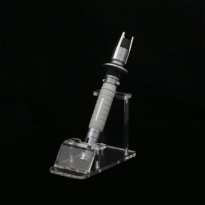 Peut inclure: Une r&eacute;plique de sabre laser argent&eacute;e avec une poign&eacute;e grise et un support en acrylique transparent. Le sabre laser est sur un fond noir.