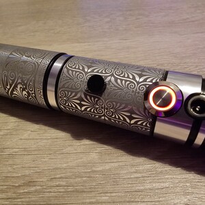 Peut inclure: Un sabre laser argent&eacute; avec des motifs floraux et g&eacute;om&eacute;triques complexes grav&eacute;s sur le manche. La poign&eacute;e comporte un bouton noir et un bouton d'alimentation circulaire rouge brillant. Le design est d&eacute;taill&eacute; et m&eacute;tallique.
