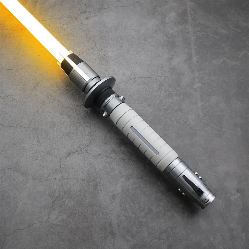 Peut inclure: Un sabre laser blanc avec une poign&eacute;e argent&eacute;e et une lame jaune. Le sabre laser est sur une surface grise.