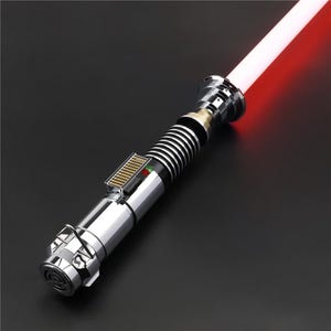 May include: A silver lightsaber with a red blade. The lightsaber has a black and silver handle with a silver button and a green and red light.