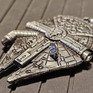 Pode incluir: Um chaveiro prateado do Millennium Falcon. O chaveiro apresenta detalhes intrincados da icônica nave espacial da franquia Star Wars. O design inclui vários painéis, aberturas e uma antena de radar central.