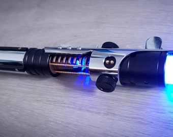 May include: A silver and black lightsaber with a blue blade. The lightsaber has a metal handle with a spring mechanism and a black knob.