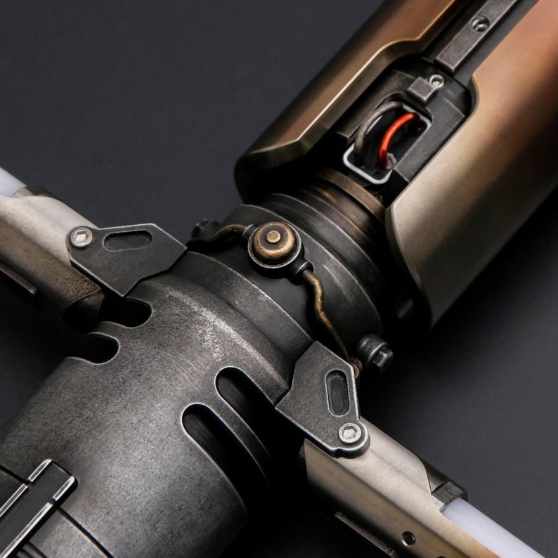 Puede incluir: Primer plano de una empu&ntilde;adura de sable de luz met&aacute;lica con un dise&ntilde;o industrial desgastado. La empu&ntilde;adura presenta una mezcla de tonos plateados, bronce y cobre, con intrincados detalles mec&aacute;nicos y un elemento rojo brillante.