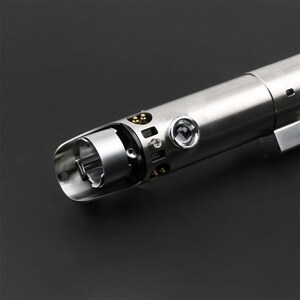 May include: A silver lightsaber hilt with a detailed design and a small, round, silver knob on the side.