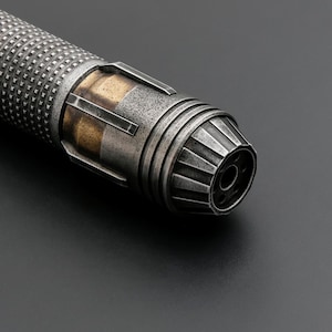 Puede incluir: Primer plano de una empu&ntilde;adura de sable de luz met&aacute;lica con un dise&ntilde;o industrial desgastado. La empu&ntilde;adura presenta un agarre texturizado, detalles dorados y un emisor detallado. La combinaci&oacute;n de colores general es plateado y dorado.