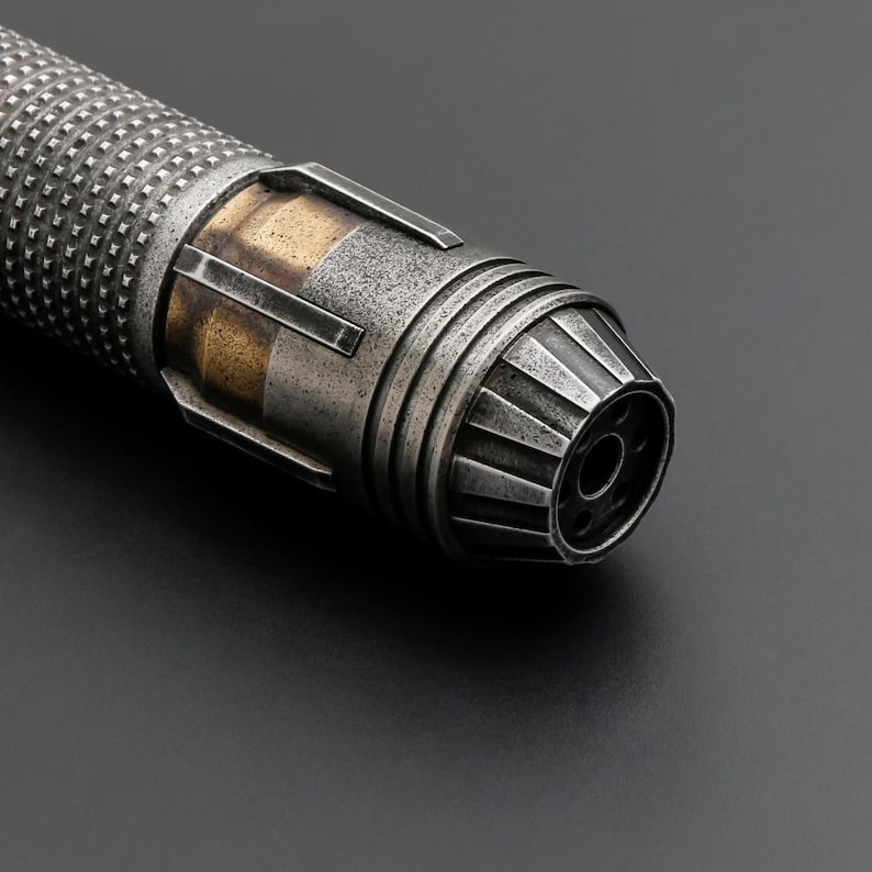 Puede incluir: Primer plano de una empu&ntilde;adura de sable de luz met&aacute;lica con un dise&ntilde;o industrial desgastado. La empu&ntilde;adura presenta un agarre texturizado, detalles dorados y un emisor detallado. La combinaci&oacute;n de colores general es plateado y dorado.