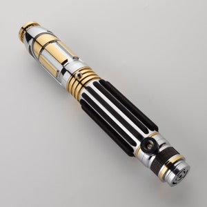 Puede incluir: Un sable de luz plateado y dorado con un agarre negro y un dise&ntilde;o texturizado. El sable de luz tiene un peque&ntilde;o bot&oacute;n redondo plateado en el lateral.