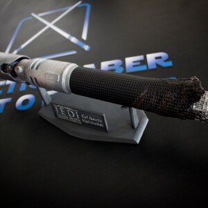 May include: A black and silver lightsaber with a burnt, damaged end. The lightsaber is on a gray stand with the text "Jedi Cal Kestis Lightsaber" on it.