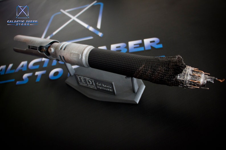 May include: A black and silver lightsaber with a burnt, damaged end. The lightsaber is on a gray stand with the text "Jedi Cal Kestis Lightsaber" on it.