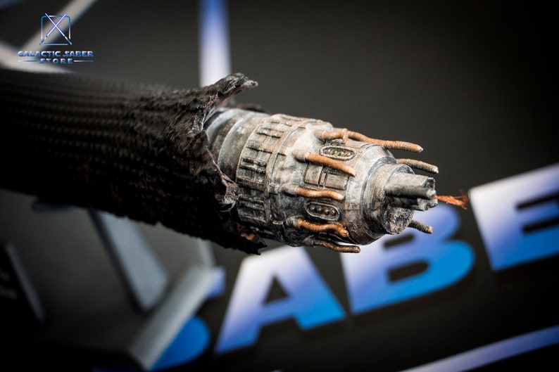 May include: A close-up of a weathered, metal lightsaber hilt with a black, textured grip. The hilt has a detailed design with multiple buttons and a small, circular window.