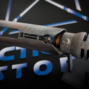 May include: A weathered, silver lightsaber hilt with a visible copper wire and internal components. The hilt is on a black stand with a "Star Wars Jedi Fallen Order" label.