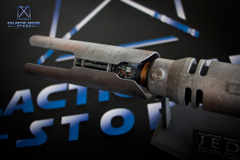 May include: A weathered, silver lightsaber hilt with a visible copper wire and internal components. The hilt is on a black stand with a "Star Wars Jedi Fallen Order" label.