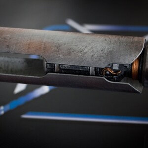 May include: A close-up of a weathered, metal lightsaber hilt with a copper-colored wire visible inside. The hilt is on a black stand.