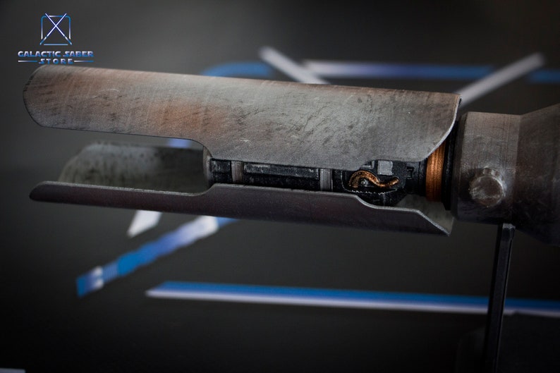 May include: A close-up of a weathered, metal lightsaber hilt with a copper-colored wire visible inside. The hilt is on a black stand.