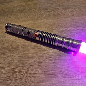 Peut inclure: Un sabre laser argent&eacute; avec des motifs grav&eacute;s complexes et une lame violette lumineuse. Le manche pr&eacute;sente une poign&eacute;e textur&eacute;e, un bouton d'activation rouge et une s&eacute;rie d'anneaux noirs et argent&eacute;s. La lame &eacute;met une lumi&egrave;re violette vive.