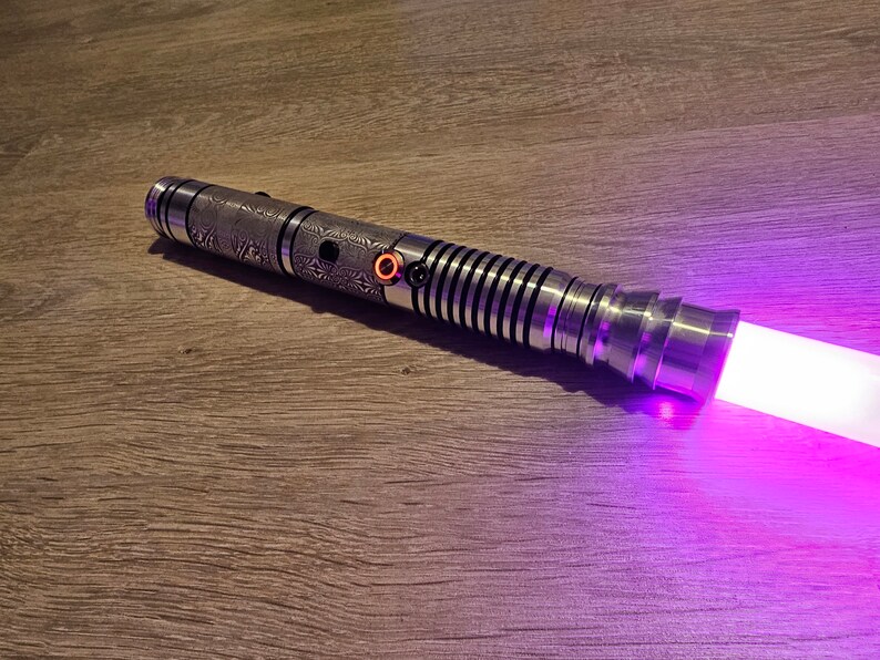 Peut inclure: Un sabre laser argent&eacute; avec des motifs grav&eacute;s complexes et une lame violette lumineuse. Le manche pr&eacute;sente une poign&eacute;e textur&eacute;e, un bouton d'activation rouge et une s&eacute;rie d'anneaux noirs et argent&eacute;s. La lame &eacute;met une lumi&egrave;re violette vive.