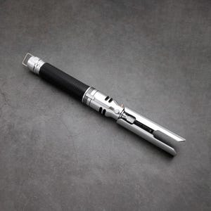 May include: A silver and black lightsaber with a black handle and a silver blade. The blade is extended and the lightsaber is lying on a grey surface.