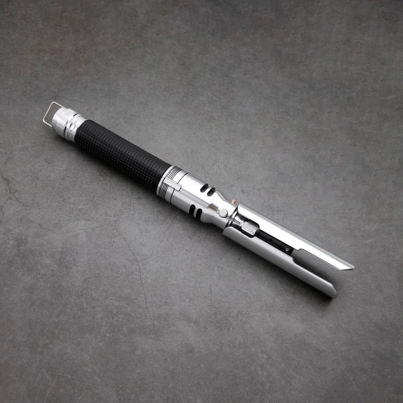 May include: A silver and black lightsaber with a black handle and a silver blade. The blade is extended and the lightsaber is lying on a grey surface.