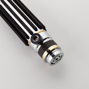 Puede incluir: Un sable de luz negro y plateado con un acento dorado y un agarre marr&oacute;n texturizado. El sable de luz tiene un mango negro y plateado con un anillo dorado alrededor del medio. El sable de luz tiene una hoja negra y plateada con una punta dorada.