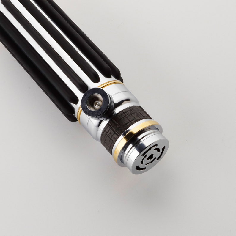 Puede incluir: Un sable de luz negro y plateado con un acento dorado y un agarre marr&oacute;n texturizado. El sable de luz tiene un mango negro y plateado con un anillo dorado alrededor del medio. El sable de luz tiene una hoja negra y plateada con una punta dorada.