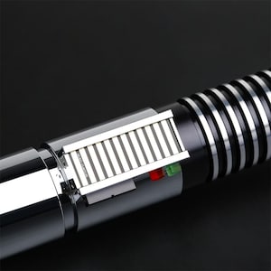 May include: A silver and black lightsaber with a silver metal panel and a green and red light. The lightsaber has a black handle with a silver ring.