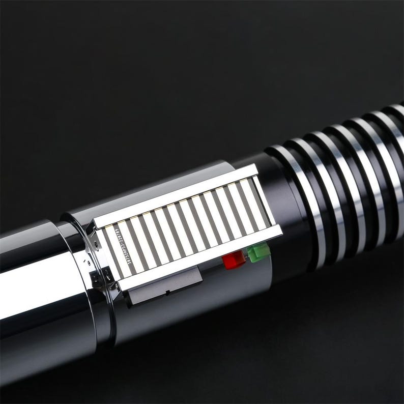 May include: A silver and black lightsaber with a silver metal panel and a green and red light. The lightsaber has a black handle with a silver ring.