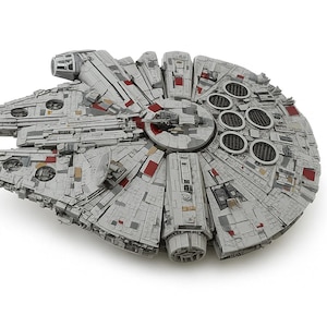 May include: A detailed model of the Millennium Falcon starship, a fictional spacecraft from the Star Wars franchise. The model is primarily gray with red and black accents, featuring intricate paneling and circular details. The ship is set against a white background.
