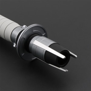 Peut inclure: Gros plan sur une poign&eacute;e de sabre laser argent&eacute;e et noire avec une poign&eacute;e blanche. La poign&eacute;e a une surface textur&eacute;e et un &eacute;metteur noir avec un anneau argent&eacute; autour.