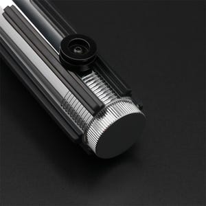 Puede incluir: Primer plano del extremo de una empu&ntilde;adura de sable l&aacute;ser negra con una tapa de metal plateado y una perilla negra.