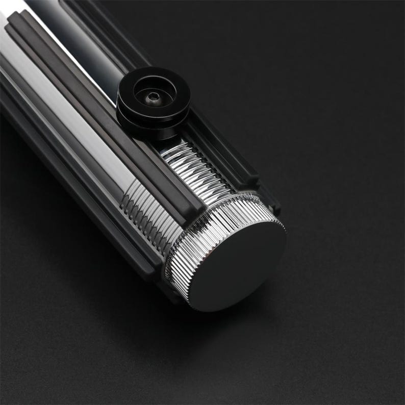 Puede incluir: Primer plano del extremo de una empu&ntilde;adura de sable l&aacute;ser negra con una tapa de metal plateado y una perilla negra.