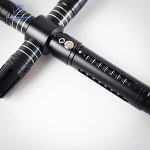 Puede incluir: Un sable de luz negro con guarda cruzada y detalles plateados. El mango tiene un bot&oacute;n y aberturas tipo ranura. El dise&ntilde;o incluye un emisor en forma de cruz. El logotipo de Galactic Saber Store es visible.