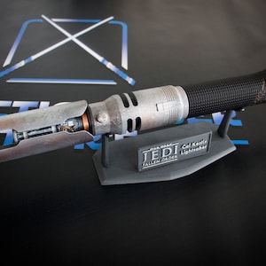 May include: A silver and black lightsaber prop with a weathered finish. The lightsaber is on a black stand with the text "STAR WARS JEDI FALLEN ORDER Cal Kestis Lightsaber".